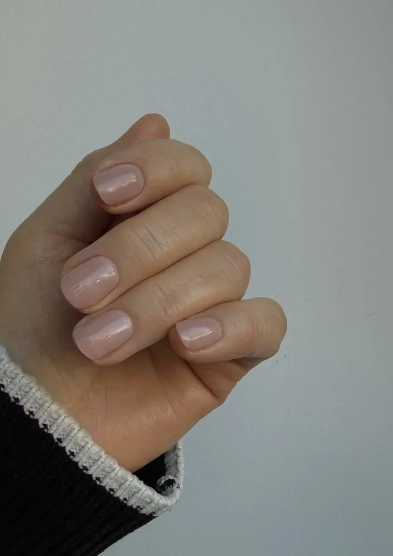 Hand with a glossy nude manicure against a pale wall